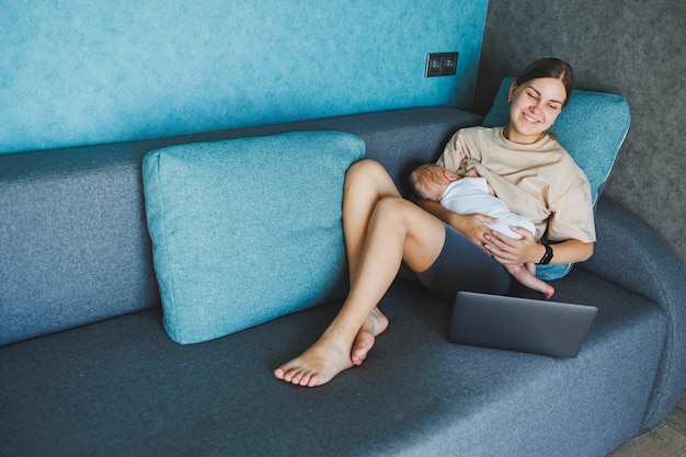 Une jeune mère souriante allaite son bébé et travaille à la maison avec son bébé sur le canapé tout en écrivant un e-mail allaitant le bébé Une jeune femme travaille à la famille