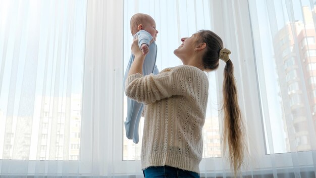 Photo jeune mère tenant son petit fils contre une grande fenêtre à la maison concept de bonheur familial
