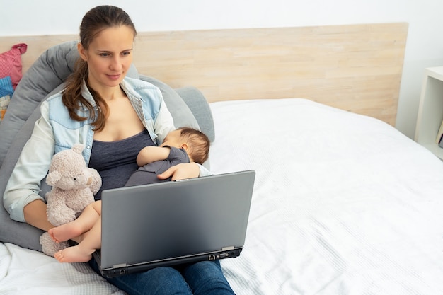 Jeune mère travaillant et utilisant un ordinateur portable avec un bébé dans ses bras et assise sur un canapé à l'intérieur