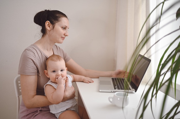 Jeune mère travaille à étudier à la maison avec un ordinateur portable, petit bébé mignon sur les genoux.