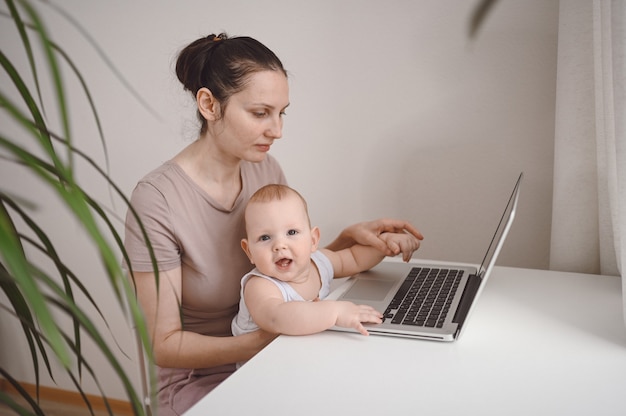 Jeune mère travaille à étudier à la maison avec un ordinateur portable, petit bébé mignon sur les genoux.