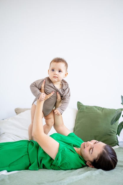 Une jeune mère en vêtements verts joue avec un petit garçon sur un lit dans une chambre lumineuse le soulevant en l'étreignant et l'embrassant une maternité heureuse