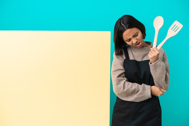 Jeune métisse cuisinier femme avec une grande pancarte isolée sur fond bleu avec douleur dans le coude