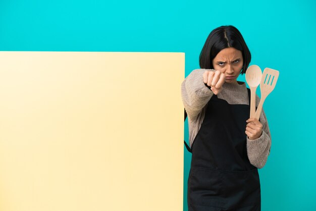 Jeune métisse cuisinier femme avec une grande pancarte isolée sur fond bleu avec geste de combat