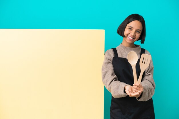 Jeune métisse cuisinier femme avec une grande pancarte isolée sur fond bleu holding copyspace imaginaire sur la paume pour insérer une annonce