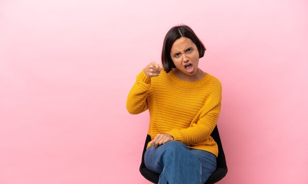 Jeune métisse femme assise sur une chaise isolée sur fond rose frustré et pointant vers l'avant