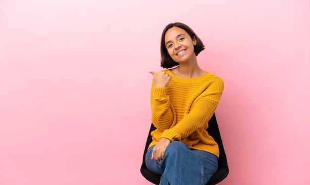 Jeune métisse femme assise sur une chaise isolée sur fond rose pointant vers le côté pour présenter un produit