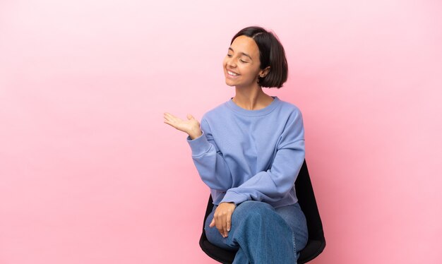 Jeune métisse femme assise sur une chaise isolée sur fond rose présentant une idée tout en regardant en souriant vers