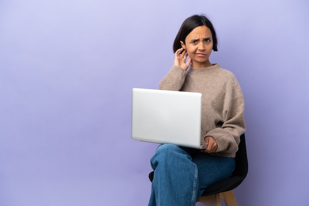 Jeune métisse femme assise sur une chaise avec ordinateur portable isolé sur fond violet ayant des doutes