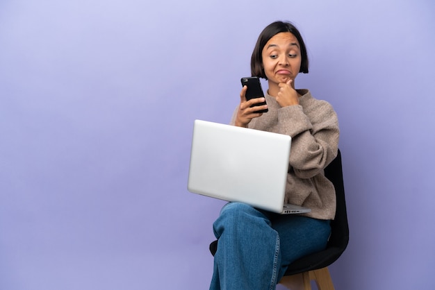 Jeune métisse femme assise sur une chaise avec ordinateur portable isolé sur fond violet penser et envoyer un message