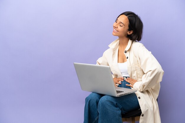 Jeune métisse femme assise sur une chaise avec ordinateur portable isolé sur fond violet posant avec les bras à la hanche et souriant