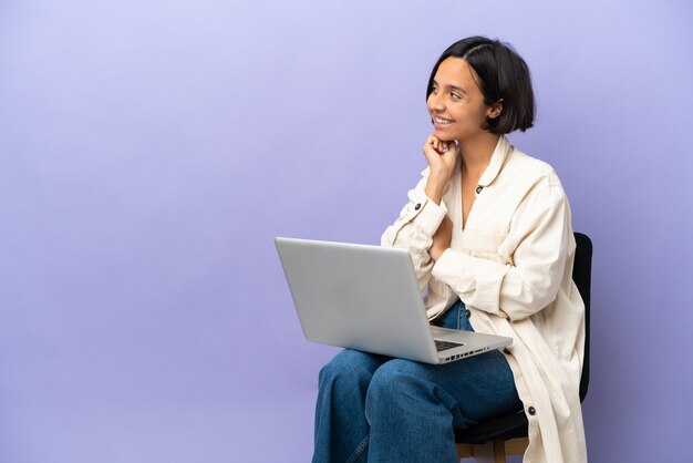 Jeune métisse femme assise sur une chaise avec ordinateur portable isolé sur fond violet regardant en souriant