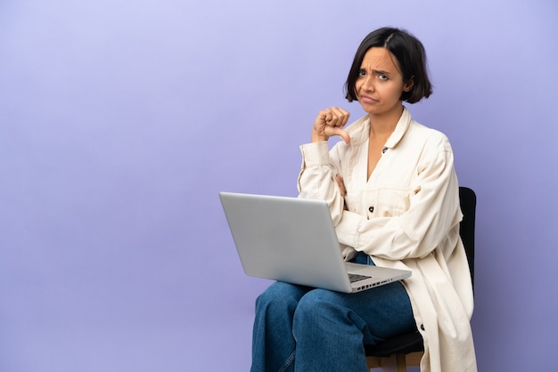 Jeune métisse femme assise sur une chaise avec un ordinateur portable isolé sur un mur violet montrant le pouce vers le bas avec une expression négative