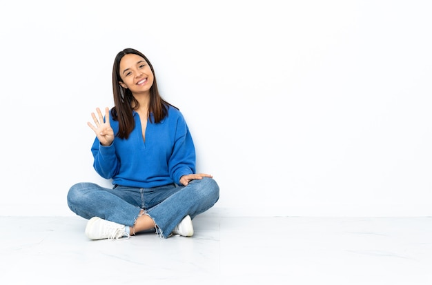 Jeune métisse femme assise sur le sol isolé sur fond blanc heureux et comptant quatre avec les doigts