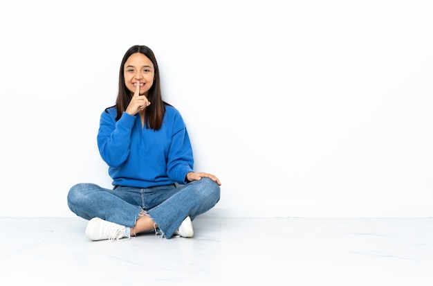 Jeune métisse femme assise sur le sol isolé sur fond blanc montrant un signe de silence geste mettant le doigt dans la bouche