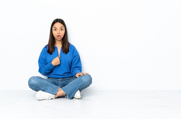 Jeune métisse femme assise sur le sol isolé sur mur blanc pointant vers soi