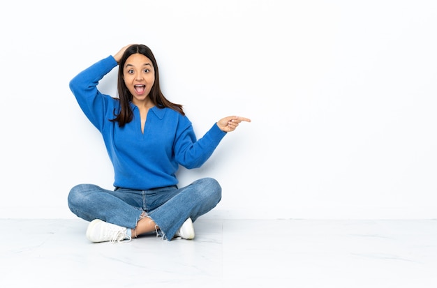 Jeune métisse femme assise sur le sol isolé sur mur blanc surpris et pointant le doigt sur le côté