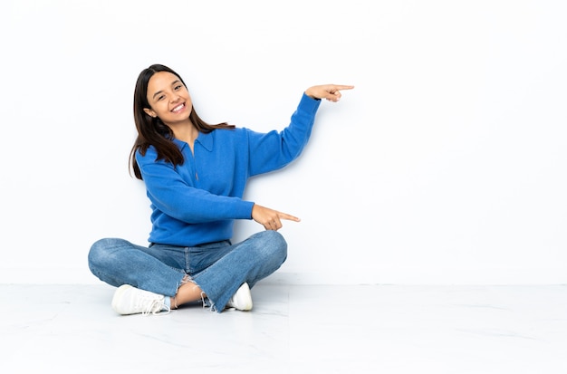 Jeune métisse femme assise sur le sol sur le mur blanc, pointant le doigt sur le côté et présentant un produit