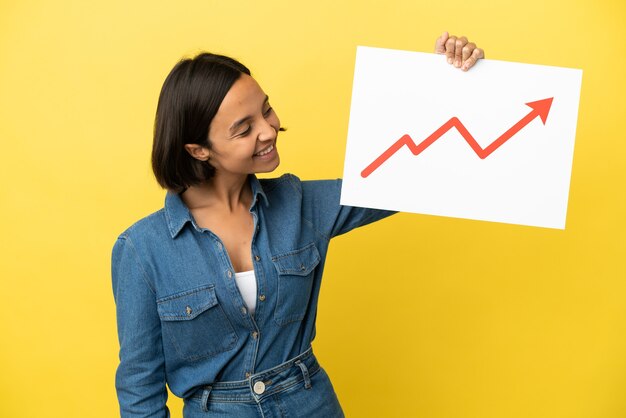 Photo jeune métisse femme isolée sur fond jaune tenant une pancarte avec un symbole de flèche de statistiques de plus en plus avec une expression heureuse