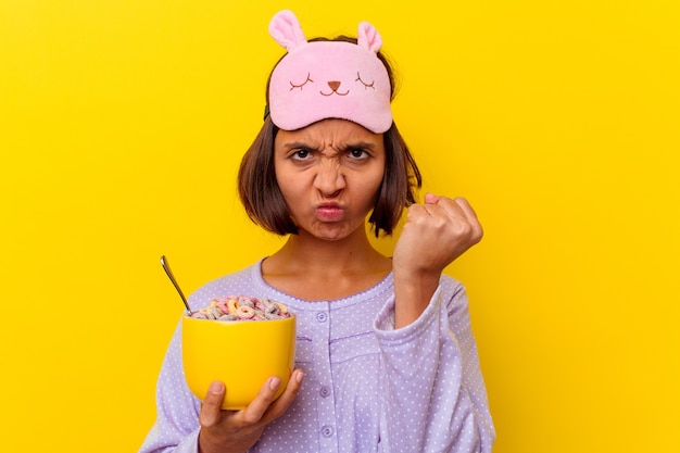 Jeune métisse femme mangeant des céréales portant un pijama isolé sur fond jaune montrant le poing à la caméra, expression faciale agressive.