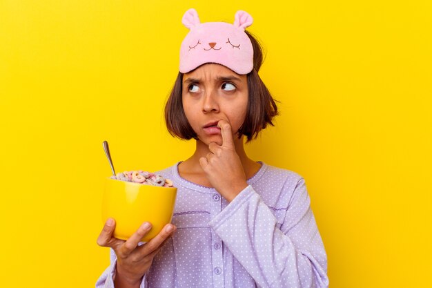 Jeune métisse femme mangeant des céréales portant un pijama isolé sur un mur jaune détendu penser à quelque chose en regardant un espace de copie.