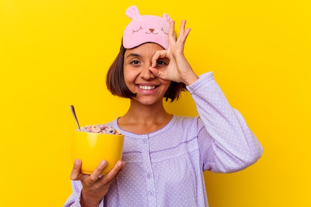 Jeune métisse femme mangeant des céréales portant un pijama isolé sur un mur jaune excité en gardant le geste ok sur les yeux.