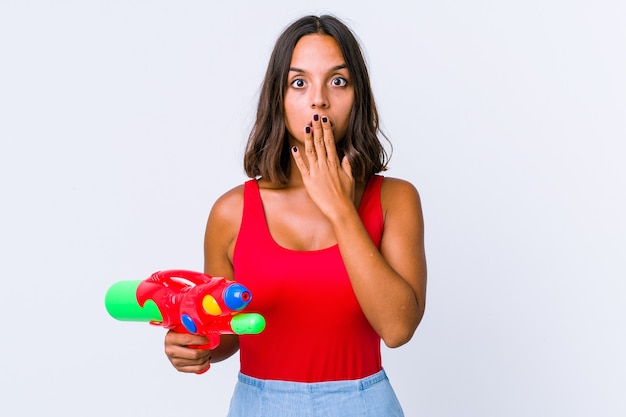 Jeune métisse femme tenant un pistolet à eau isolé choqué, couvrant la bouche avec les mains, impatient de découvrir quelque chose de nouveau.