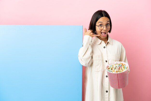 Jeune métisse femme tenant des pop-corn avec une grande bannière sur fond isolé faisant un geste de téléphone. Rappelez-moi signe