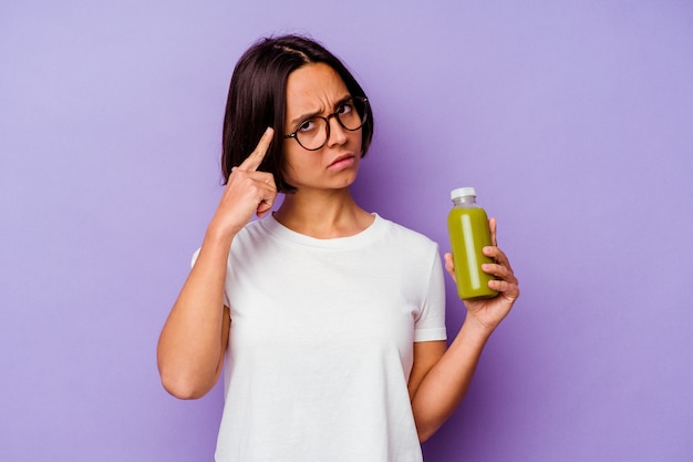 Jeune métisse femme tenant un smoothie sain isolé sur fond violet pointant le temple avec le doigt, pensant, concentré sur une tâche.