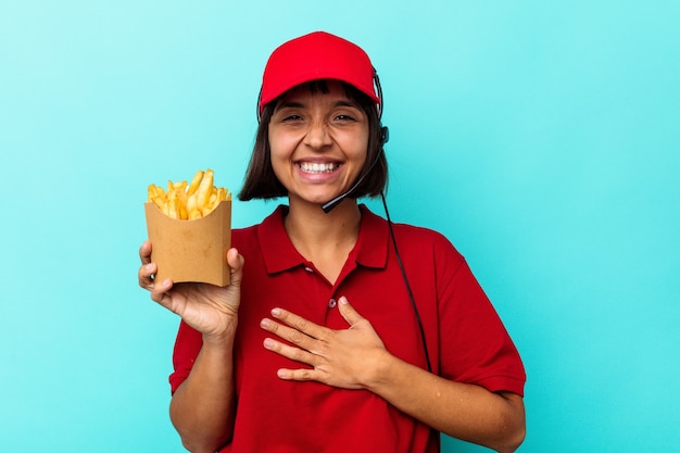 Jeune Métisse Femme Travailleur De Restauration Rapide Tenant Des Frites Isolées Sur Fond Bleu Rit Fort En Gardant La Main Sur La Poitrine.