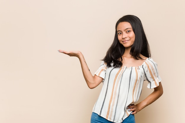 Jeune mignon adolescent chinois Jeune femme blonde portant un manteau contre un mur rose montrant un espace de copie sur une paume et tenant une autre main sur la taille.