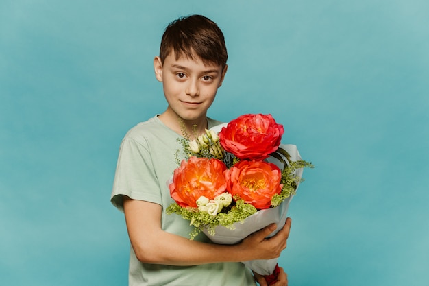 Jeune mignon garçon caucasien, sourire mignon, porte une chemise vert clair, détient des fleurs roses avec un parfum agréable, des modèles sur le mur bleu. Printemps