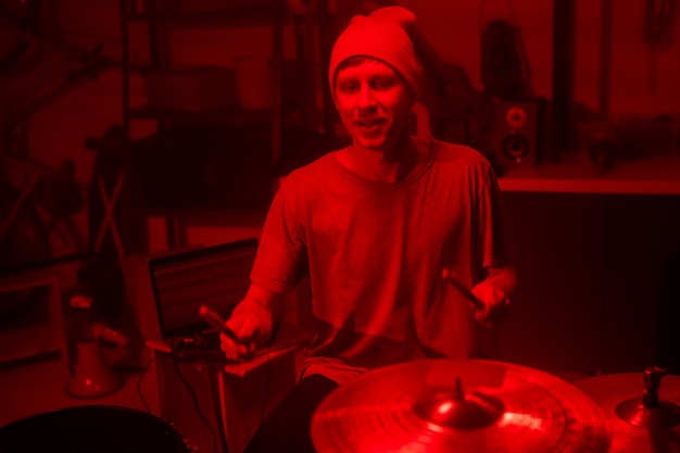 Jeune musicien en bonnet et t-shirt frappant des cymbales et des tambours avec des baguettes en position assise