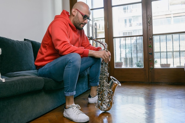 jeune musicien homme à la maison assis calme regardant vers le bas pensant tenant son saxophone