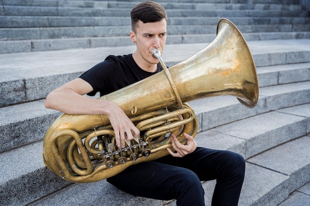 Jeune musicien de rue jouant du tuba assis sur des marches de granit