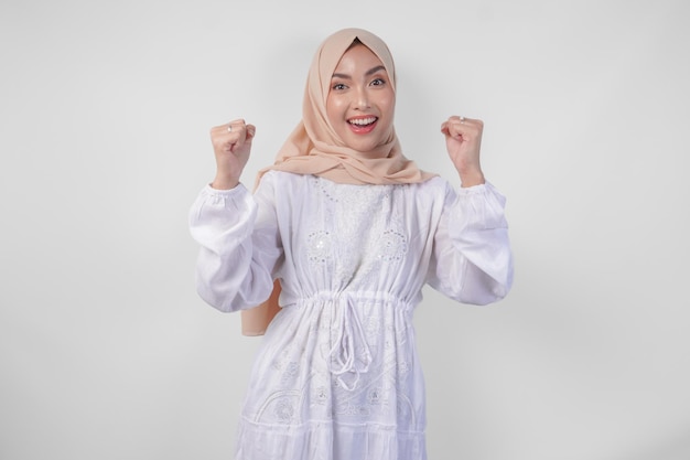 Une jeune musulmane asiatique enjouée portant un hijab et une robe blanche serrant le poing geste du vainqueur isolé sur un fond blanc concept de Ramadan et Eid Mubarak