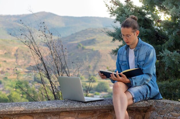 Jeune nomade numérique concentré dessinant sur son cahier avec un ordinateur portable appuyé sur le sol