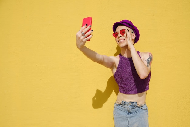 Jeune non binaire souriant et posant devant l'appareil photo du téléphone portable tout en prenant des selfies à l'extérieur contre un mur jaune.