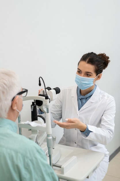 Jeune ophtalmologiste en blouse blanche et masque de protection assise près d'un équipement médical tout en allant tester la vue d'un patient masculin âgé