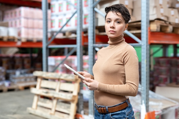 Jeune ouvrier d'entrepôt confiant avec tablette debout contre des racks