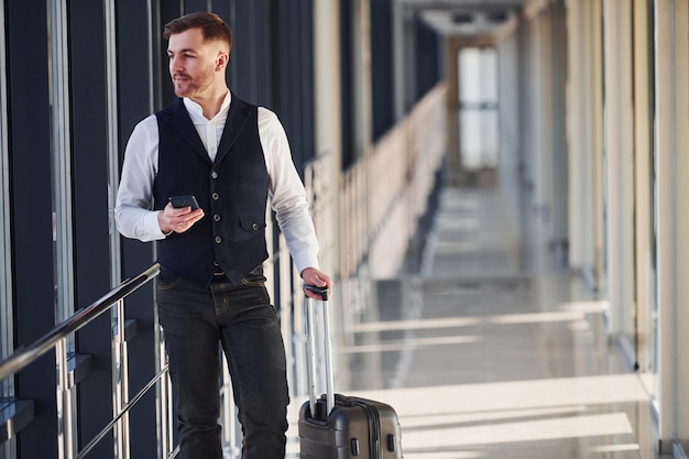 Un jeune passager masculin vêtu d'élégants vêtements formels se trouve dans le hall de l'aéroport, debout avec ses bagages et son téléphone à la main.