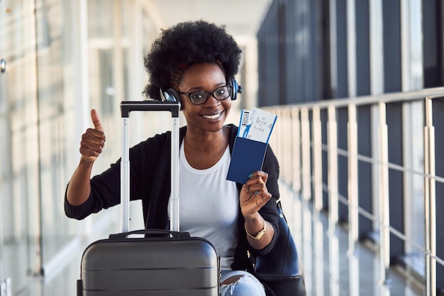 Une jeune passagère afro-américaine en tenue décontractée et casque est à l'aéroport avec des bagages.