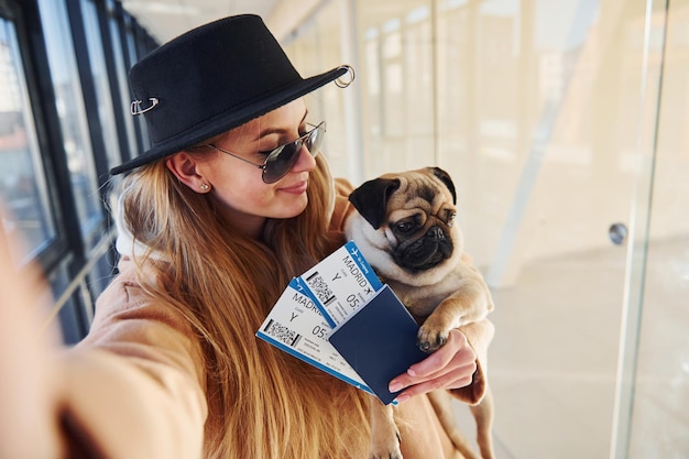 Jeune passagère dans des vêtements chauds tenant des billets et un chien mignon dans les mains dans le hall de l'aéroport.