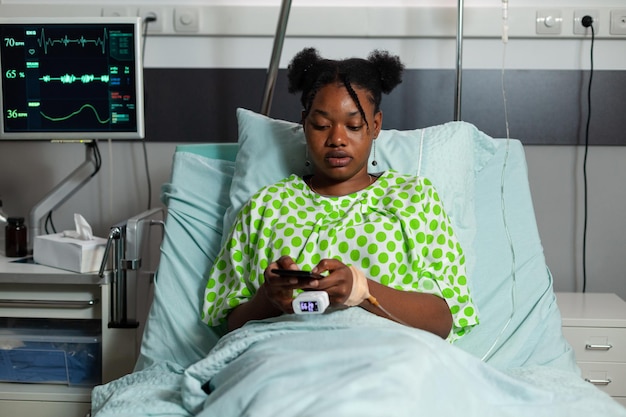 Photo jeune patient afro-américain se reposant dans son lit tenant un smartphone moderne naviguant sur internet lors d'un rendez-vous médical à l'hôpital. messagerie de femme malade avec des amis. service de médecine