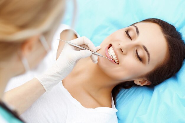 Une jeune patiente visite le cabinet du dentiste. Une belle femme aux dents blanches et droites en bonne santé assise sur un fauteuil dentaire avec la bouche ouverte pendant un examen buccal pendant que le médecin travaille aux dents.