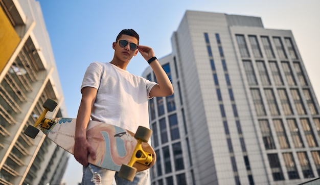 Jeune patineur américain caucasien élégant tenant un longboard à la main, dans des verres, dans le contexte des bâtiments urbains.