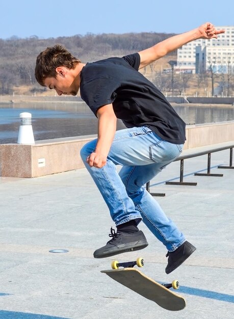 Jeune patineur russe en t-shirt noir et jean bleu sautant sur le skateboard au début du printemps