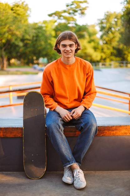 Jeune patineur souriant en pull orange et jeans regardant joyeusement à huis clos avec planche à roulettes au skatepark moderne