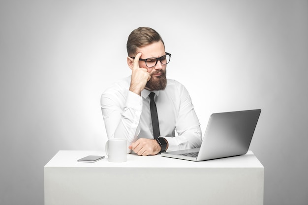 Un jeune patron barbu réfléchi en chemise blanche et cravate noire est assis au bureau sur un bureau et regarde un rapport quotidien sur un ordinateur portable, ayant une nouvelle idée et planifiant sa propre stratégie, tenant une main sur la tête.