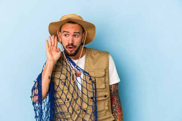 Jeune pêcheur caucasien avec des tatouages tenant un filet isolé sur fond bleu essayant d'écouter un potin.